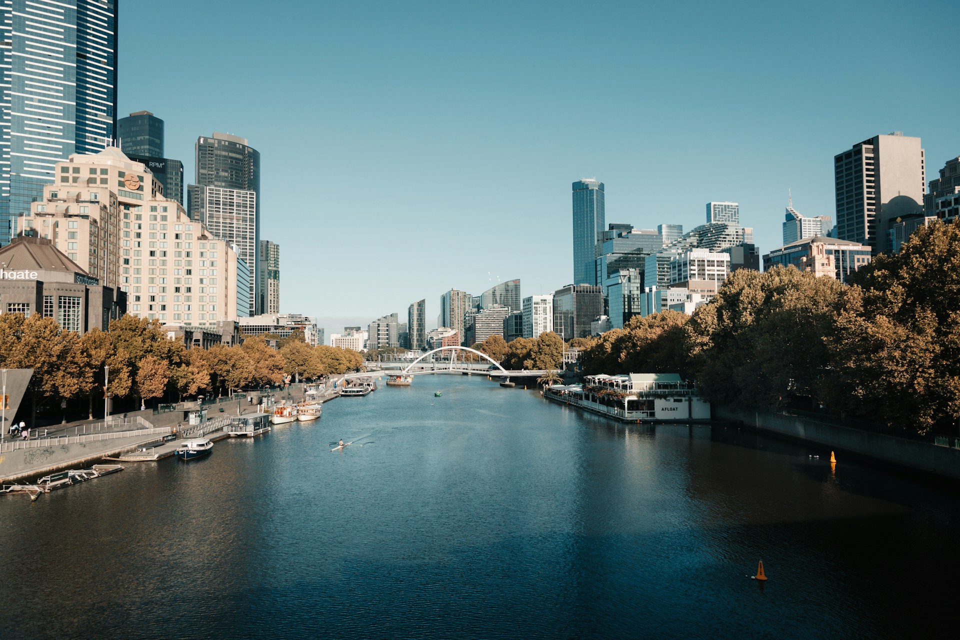 River running through a city centre, lined with trees and high-rise buildings under a clear sky