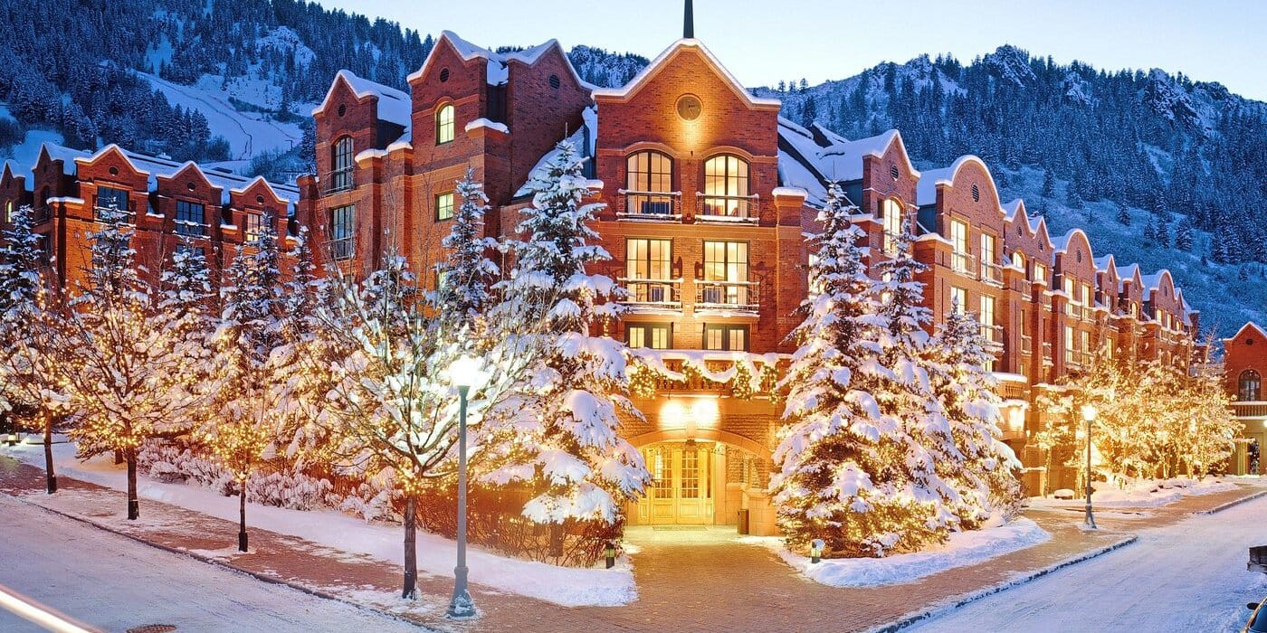 Winter view of a luxury red-brick hotel in Aspen, Colorado, surrounded by snow-covered trees and mountains.