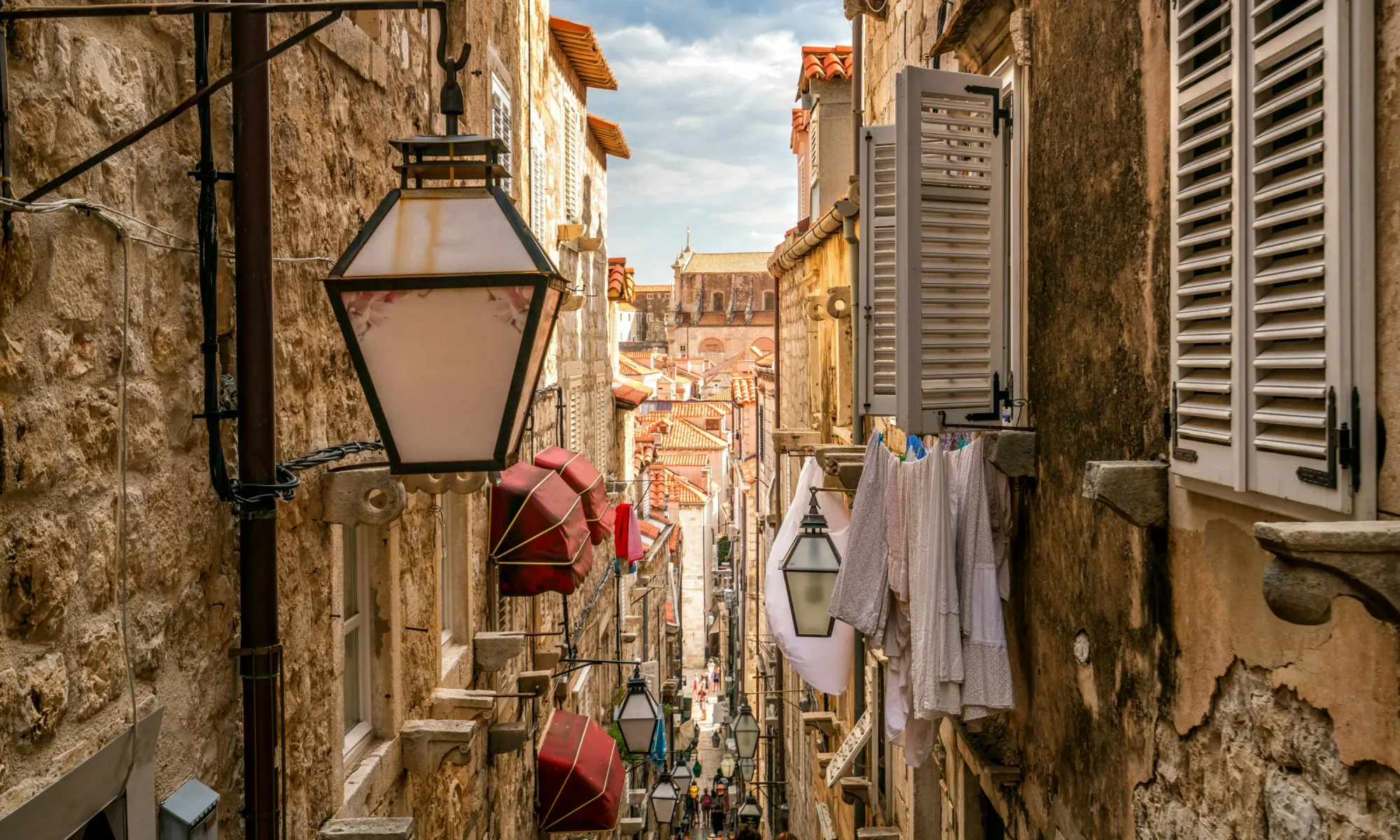 Stone alleyway in Dubrovnik’s old town with hanging lanterns, laundry drying from windows, and red-tiled rooftops visible in the distance.