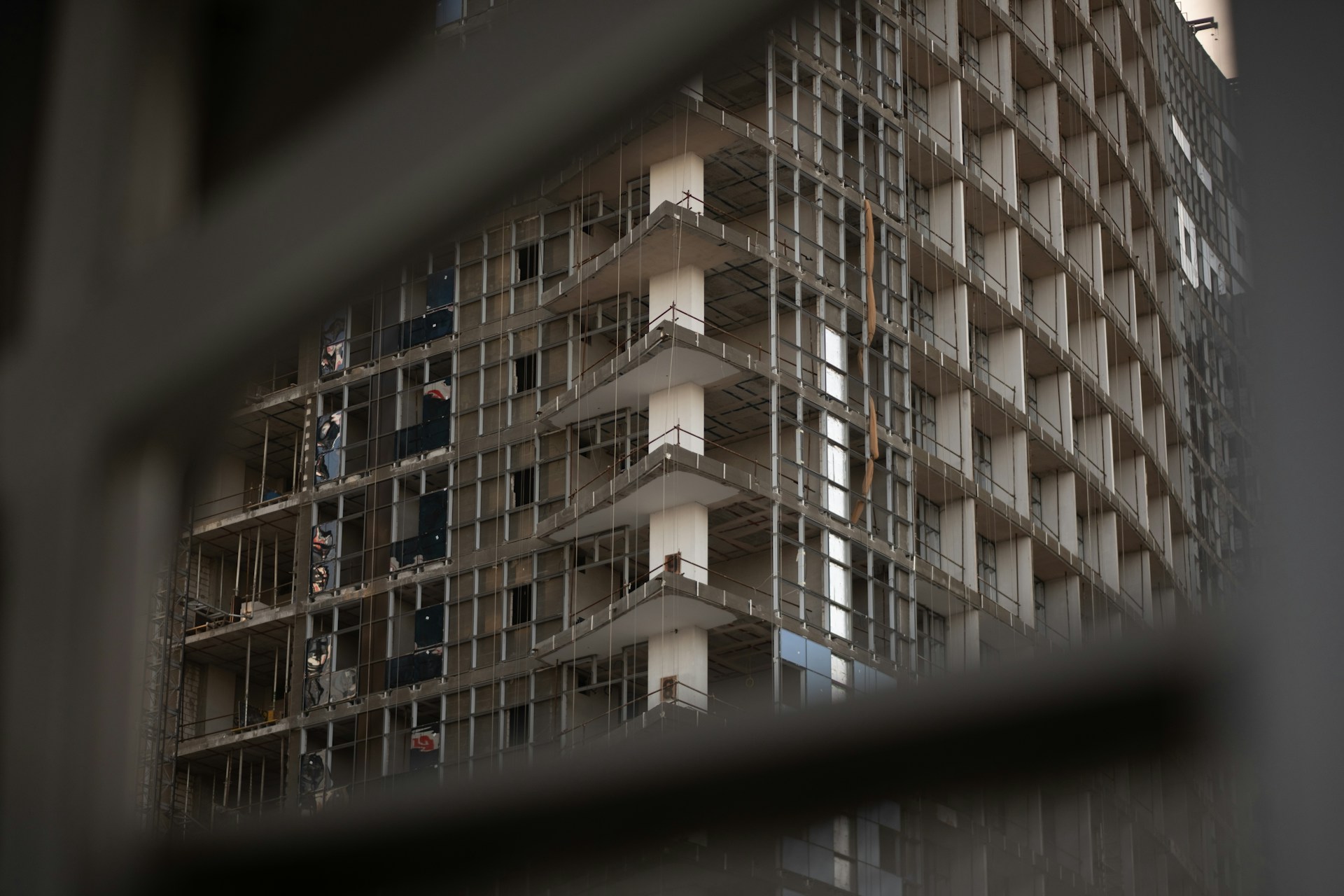 High-rise residential building under construction with unfinished facade, seen through a blurred fence